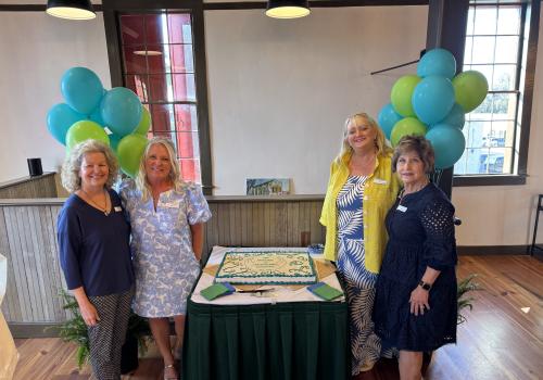 Four women smiling around a decorated cake with green and blue balloons in the background.