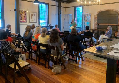 Audience seated indoors, listening to a person standing and speaking.